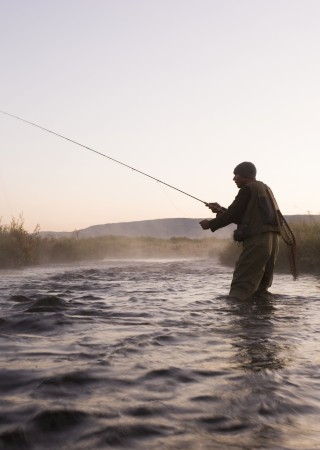 A person in outdoor gear fishes in a river at dawn or dusk, surrounded by nature and mist, with mountains in the background.
