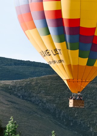 A colorful hot air balloon floats over green hills with some trees visible in the foreground and a clear sky above.
