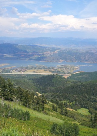 A scenic view of a lake surrounded by mountains, trees, and a partly cloudy sky in a lush, green landscape.