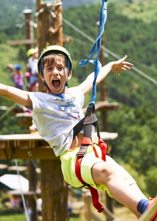 A boy is ziplining outdoors with arms spread wide, mid-air, wearing a helmet and harness, surrounded by green trees and other children.