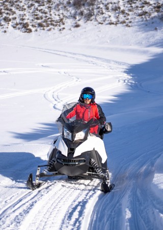 People are riding snowmobiles on a snowy landscape, with one person in the foreground and others following behind in a line.