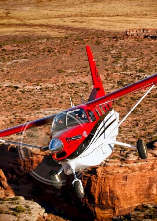 A red and white aircraft flying over a desert canyon landscape.