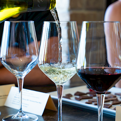 A person is pouring white wine into a glass, with other wine glasses and chocolates on a table, likely during a wine tasting event.