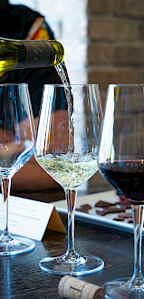 A person pours white wine into a glass, with red wine and chocolates on the table, in a cozy indoor setting.