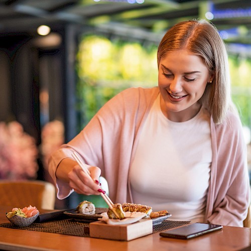 A woman is happily enjoying sushi at a restaurant, with a glass of water and a mobile phone on the table.