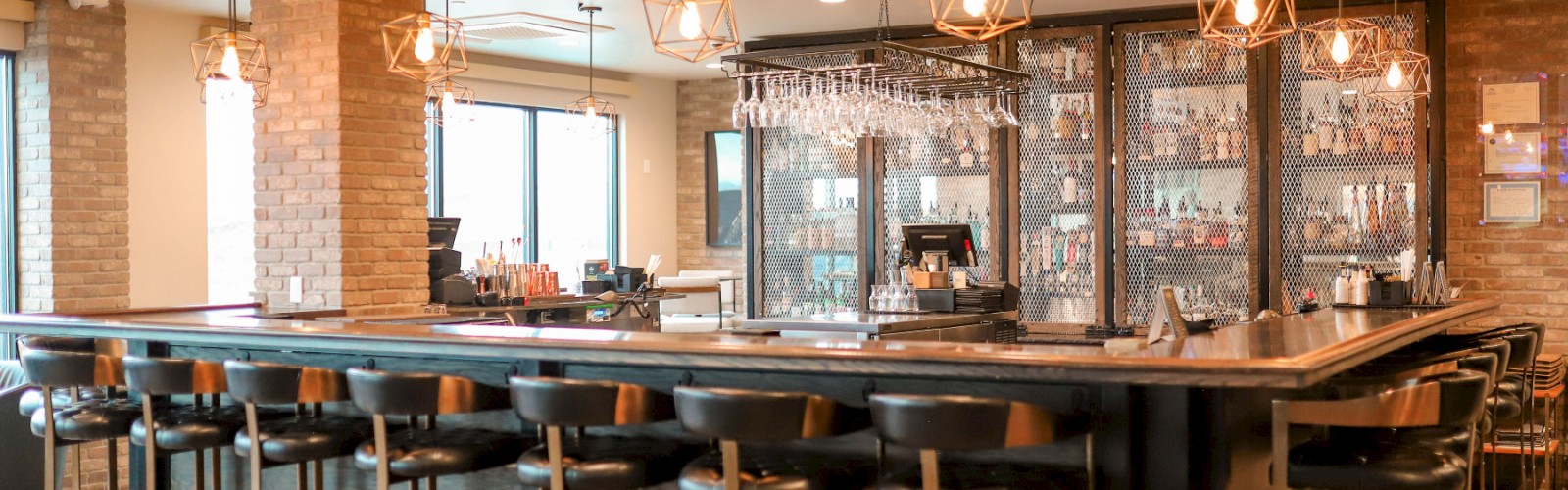 A modern bar with black seats, pendant lights, exposed brick walls, and a well-stocked back bar area.