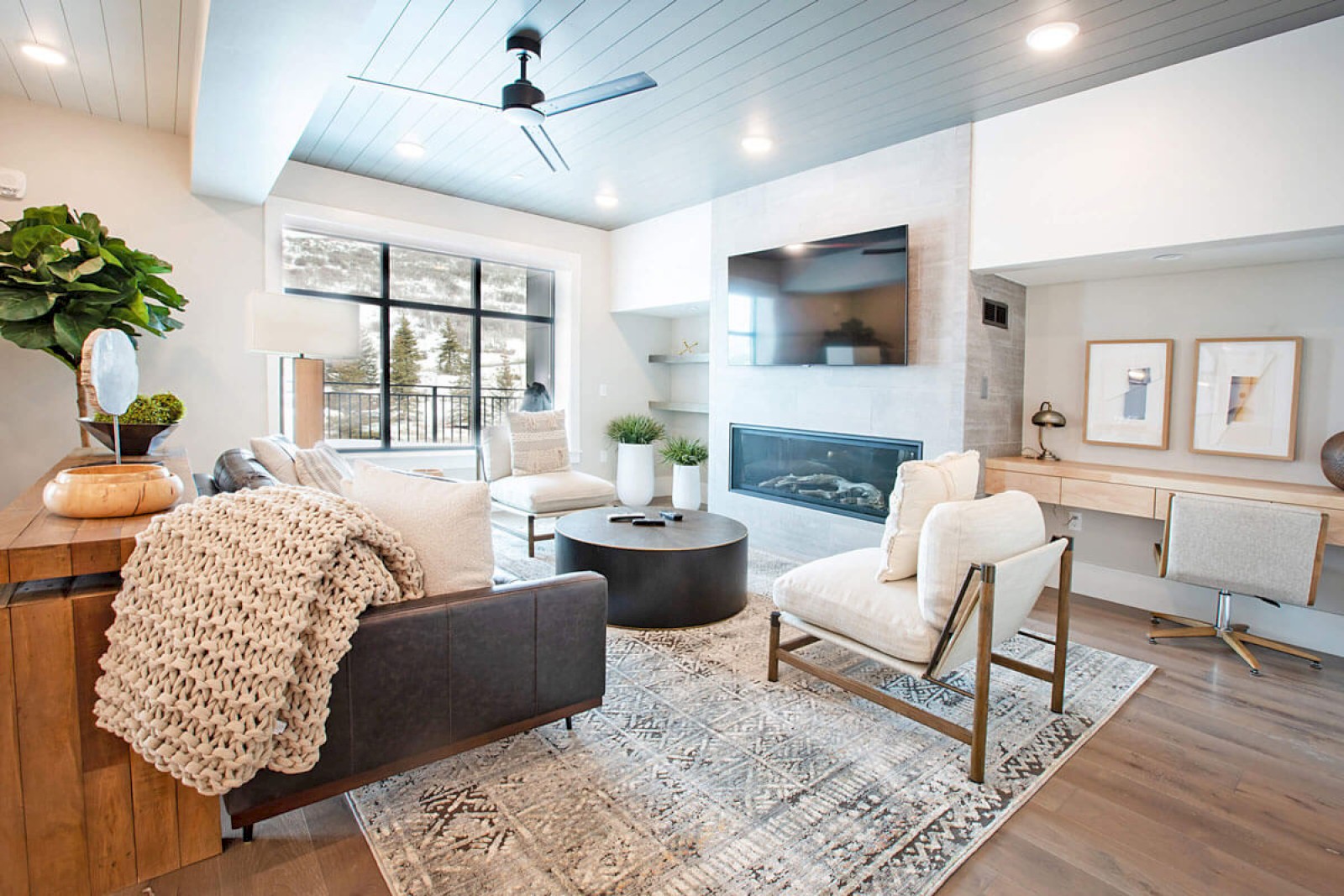 A cozy, modern living room with white walls, a large window, plush seating, a fireplace, and stylish decor.