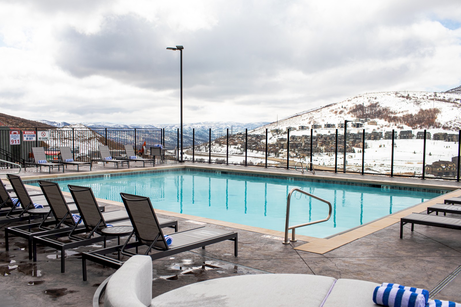 An outdoor pool area with lounge chairs, snowy mountains, and a cloudy sky in the background.
