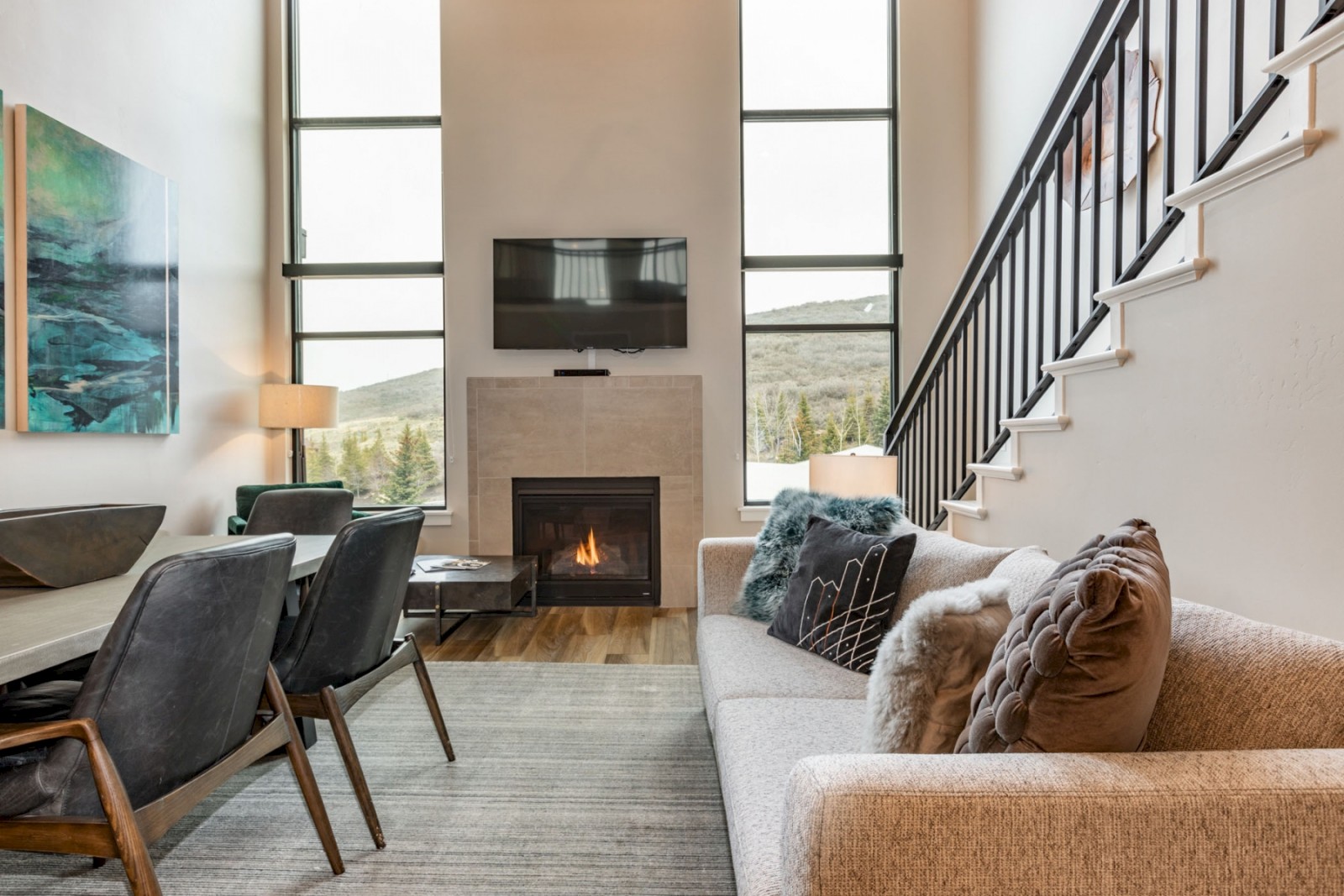 A modern living room with a fireplace, large windows showing a mountain view, and cozy furniture, including a sofa with decorative pillows.