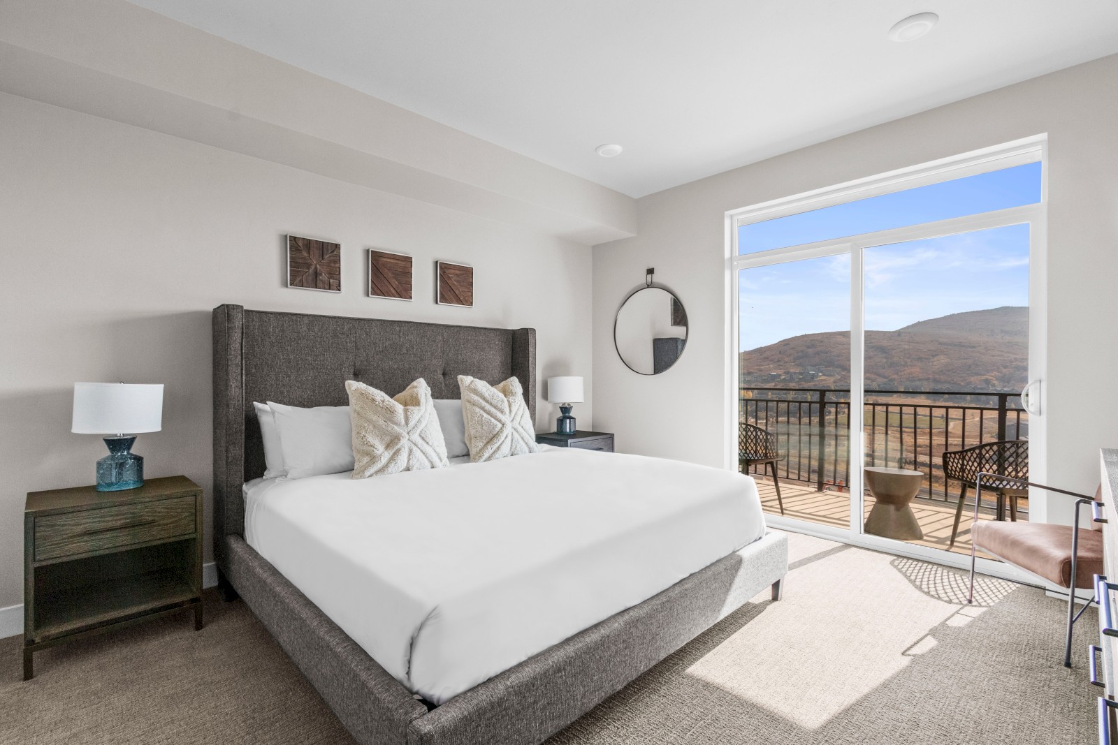 A modern bedroom with a large bed, balcony view of mountains, outdoor chairs, and minimal wall decor in a bright, airy space.