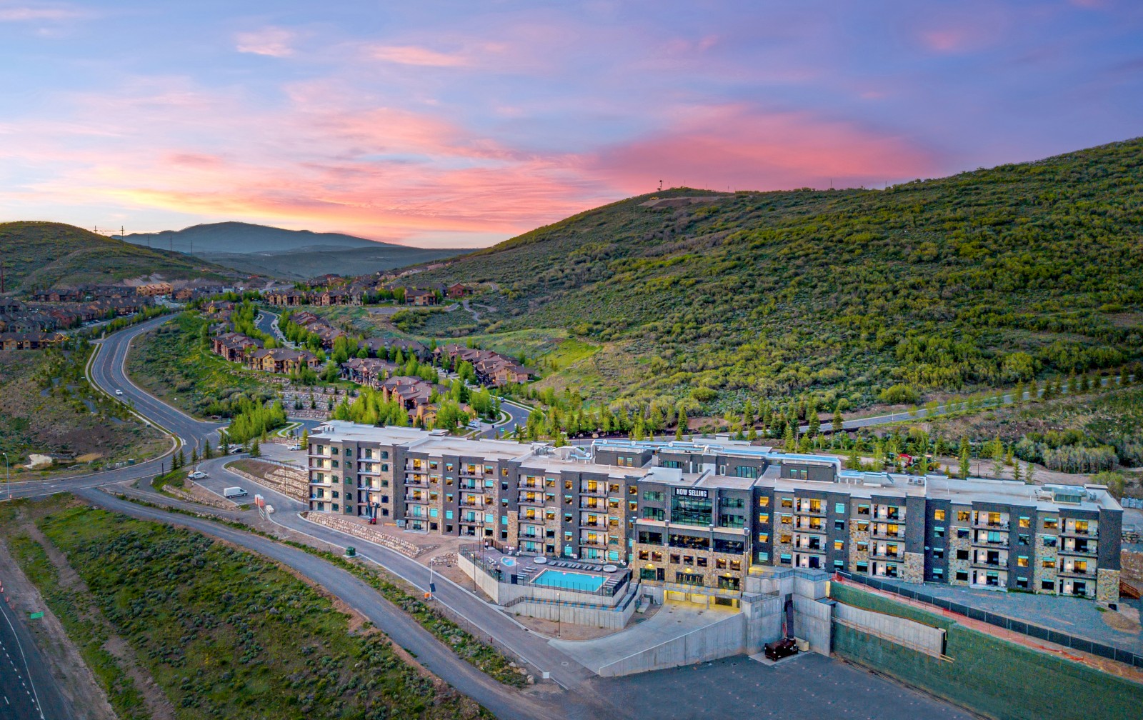 A modern apartment complex with a pool is situated in a hilly, green landscape during sunset, surrounded by roads and residential areas.