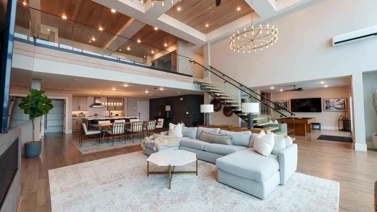 A spacious, modern open-plan living area with a staircase, high ceilings, chandeliers, and a mix of cozy and stylish furniture.