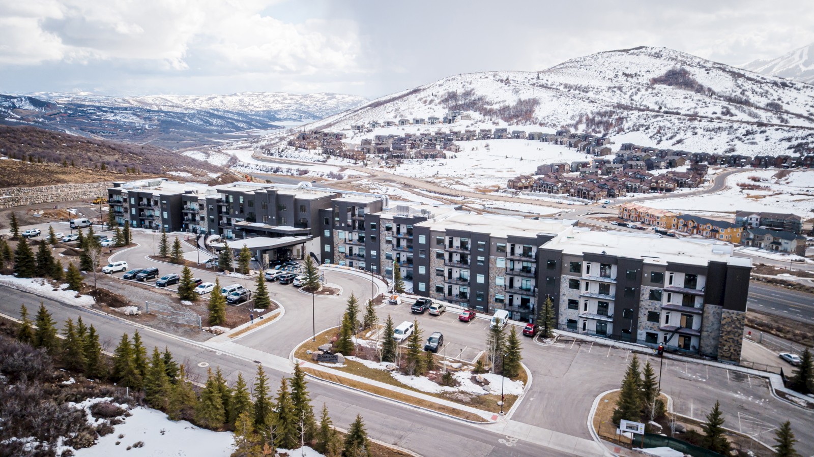 A modern apartment complex with parking lots, surrounded by snow-capped mountains and smaller buildings in a mountainous area.