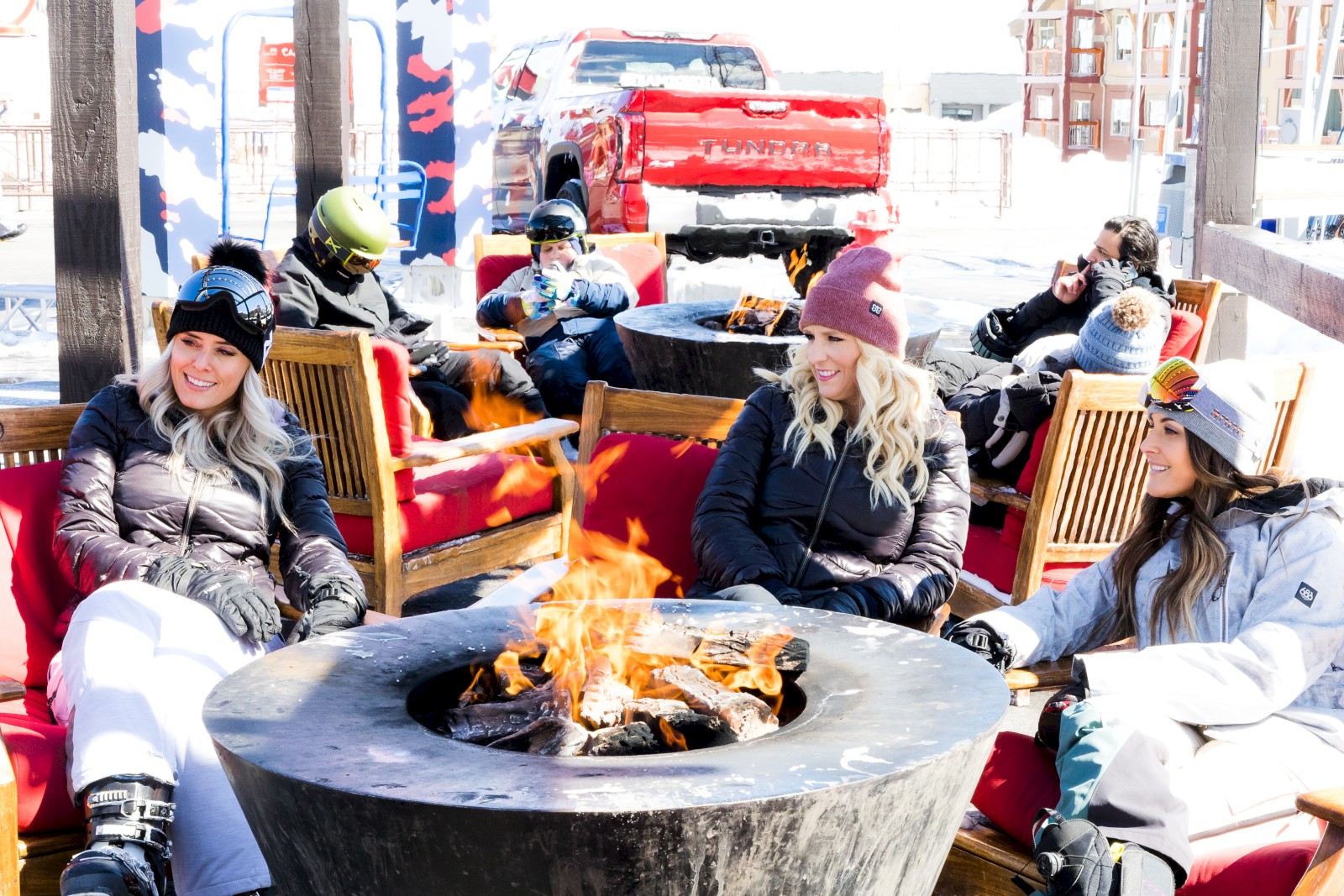 Friends enjoying a snowy day around a fire pit, dressed warmly with skis and snow gear.