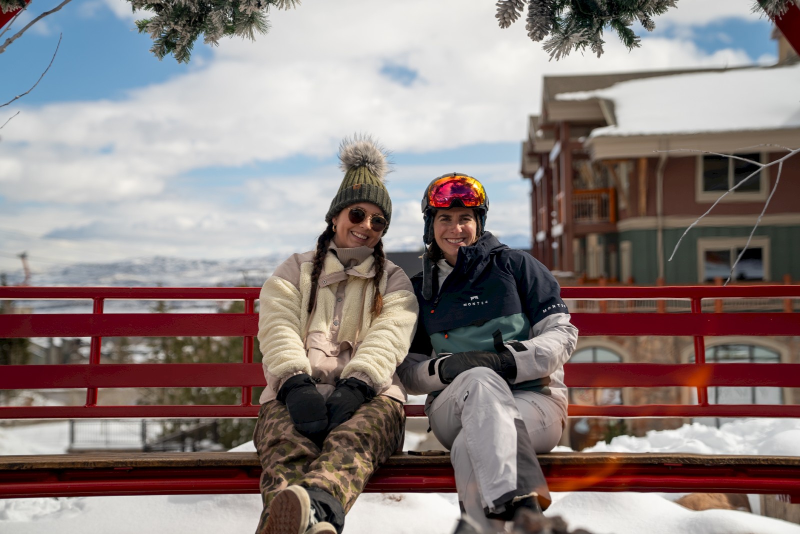 Two people are sitting on a red bench outdoors in winter, dressed warmly with snow and buildings in the background.