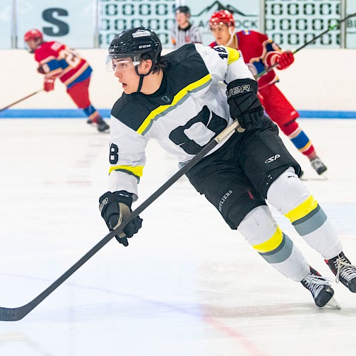 A hockey player in a black and white jersey is skating on ice during a game, with opponents in red jerseys visible in the background.