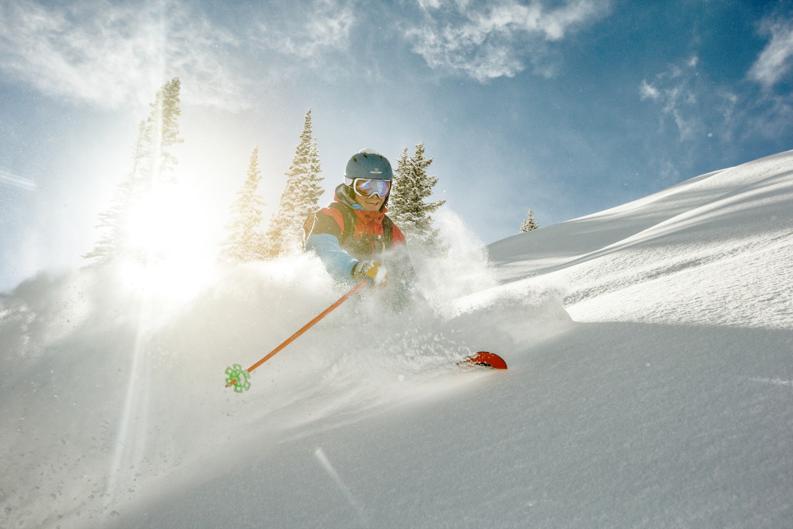 A skier happily descends a snowy mountain slope under a bright blue sky with tall pine trees nearby.