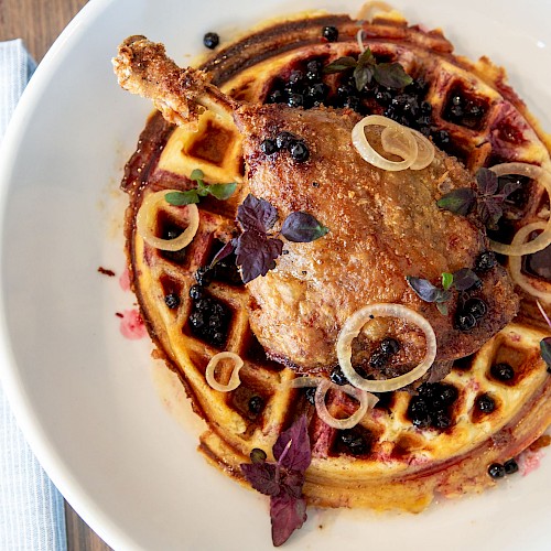 A plate with a waffle topped with a piece of fried chicken, onions, and garnished with herbs, beside a napkin and cutlery.