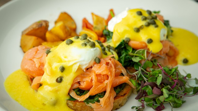 The image shows eggs Benedict with salmon, Hollandaise sauce, capers, roasted potatoes, and a side of mixed greens on a white plate.