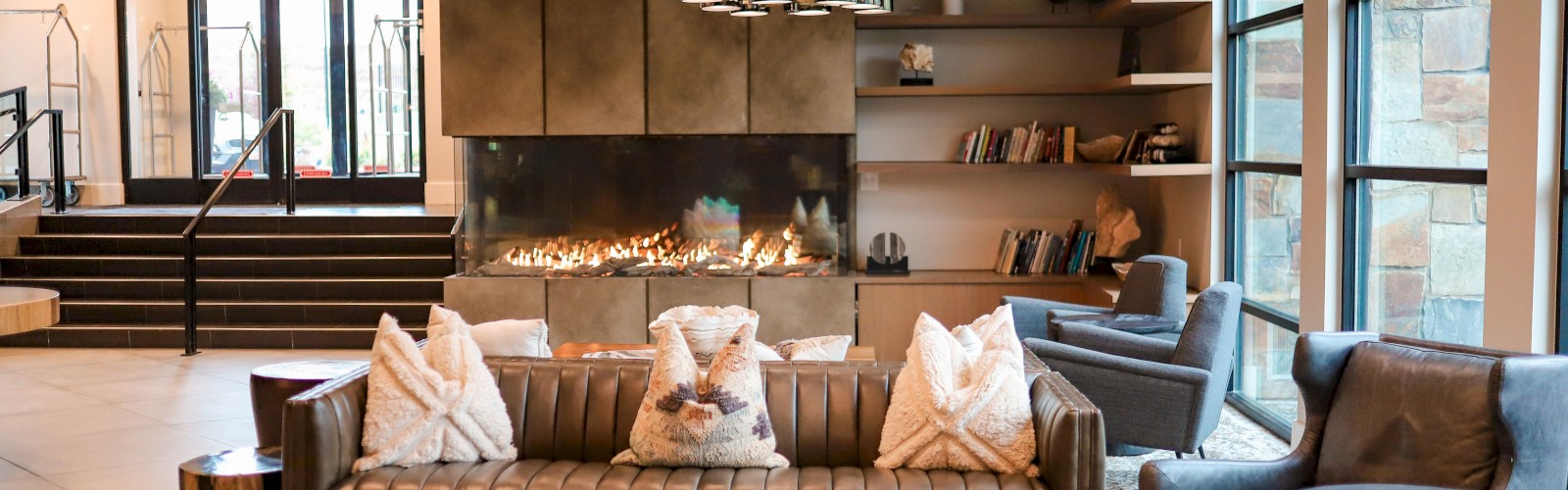 A modern living room with a large sofa, fireplace, shelves with books, and seating, featuring ample natural light from large windows.