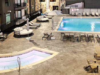 A swimming pool, hot tub, lounge chairs, umbrellas, and seating area within an apartment complex or hotel courtyard during the evening.
