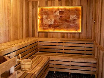 The image shows a modern sauna with wooden benches and a decorative wall panel illuminated by warm lighting. A ladle and bucket are placed on a bench.