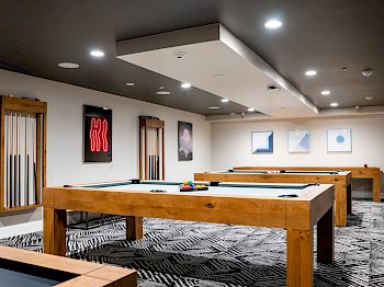 The image shows a modern room with pool tables, wall art, and cues in a rack, featuring contemporary lighting and a patterned carpet.