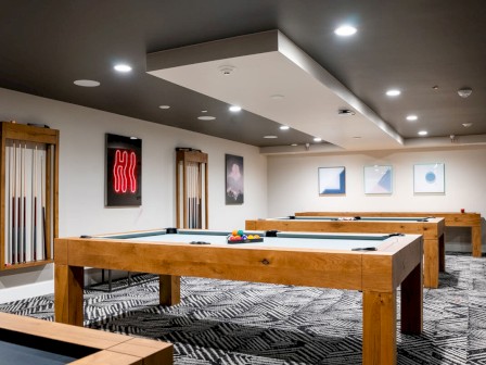 The image shows a modern room with pool tables, wall art, and cues in a rack, featuring contemporary lighting and a patterned carpet.