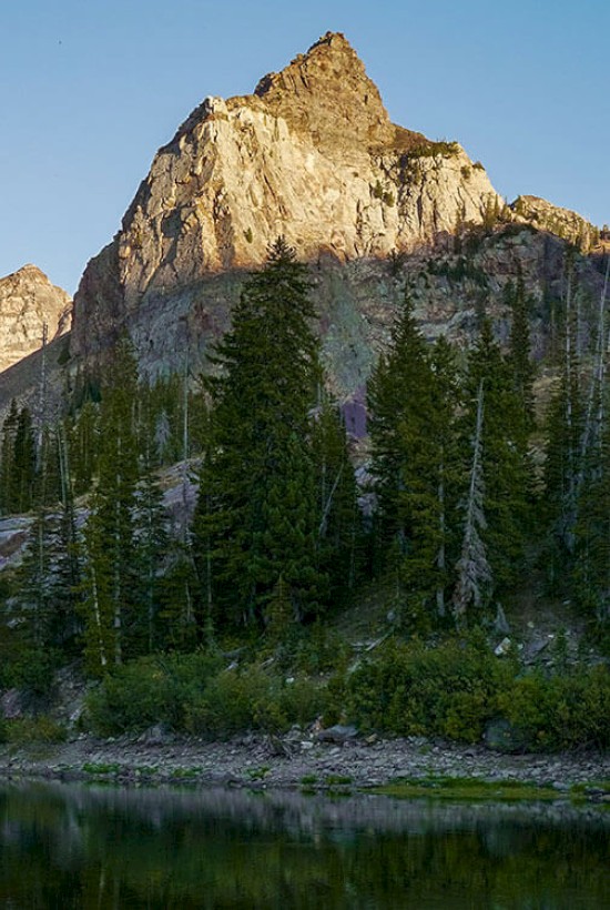A rocky mountain peak stands tall in the background, with a forest of evergreen trees and a serene lake in the foreground, under a clear sky.