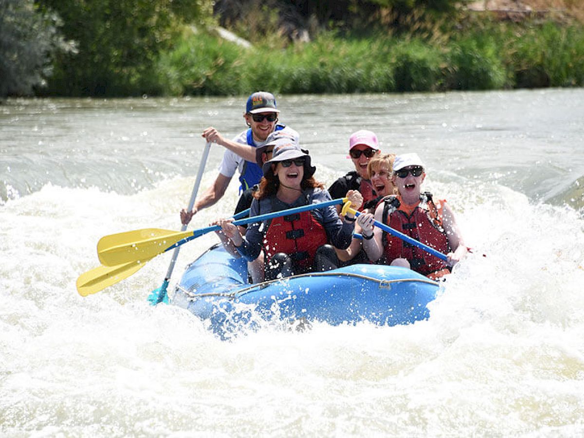 A group of people are enjoying white-water rafting in a blue raft on a flowing river, wearing life jackets and helmets, paddling through the rapids.