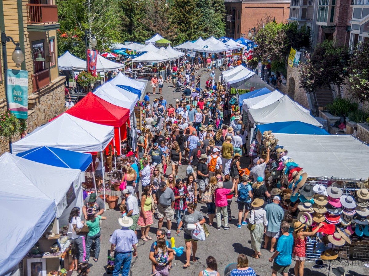 A lively outdoor market with numerous tents and shoppers exploring stalls, possibly in a town or city on a sunny day.