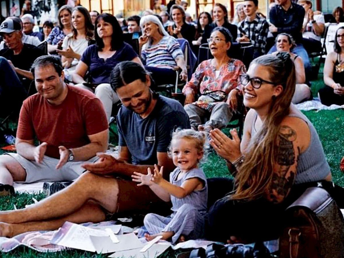 A group of people seated outdoors on a lawn, smiling and clapping, with a young child sitting on a blanket in the front.