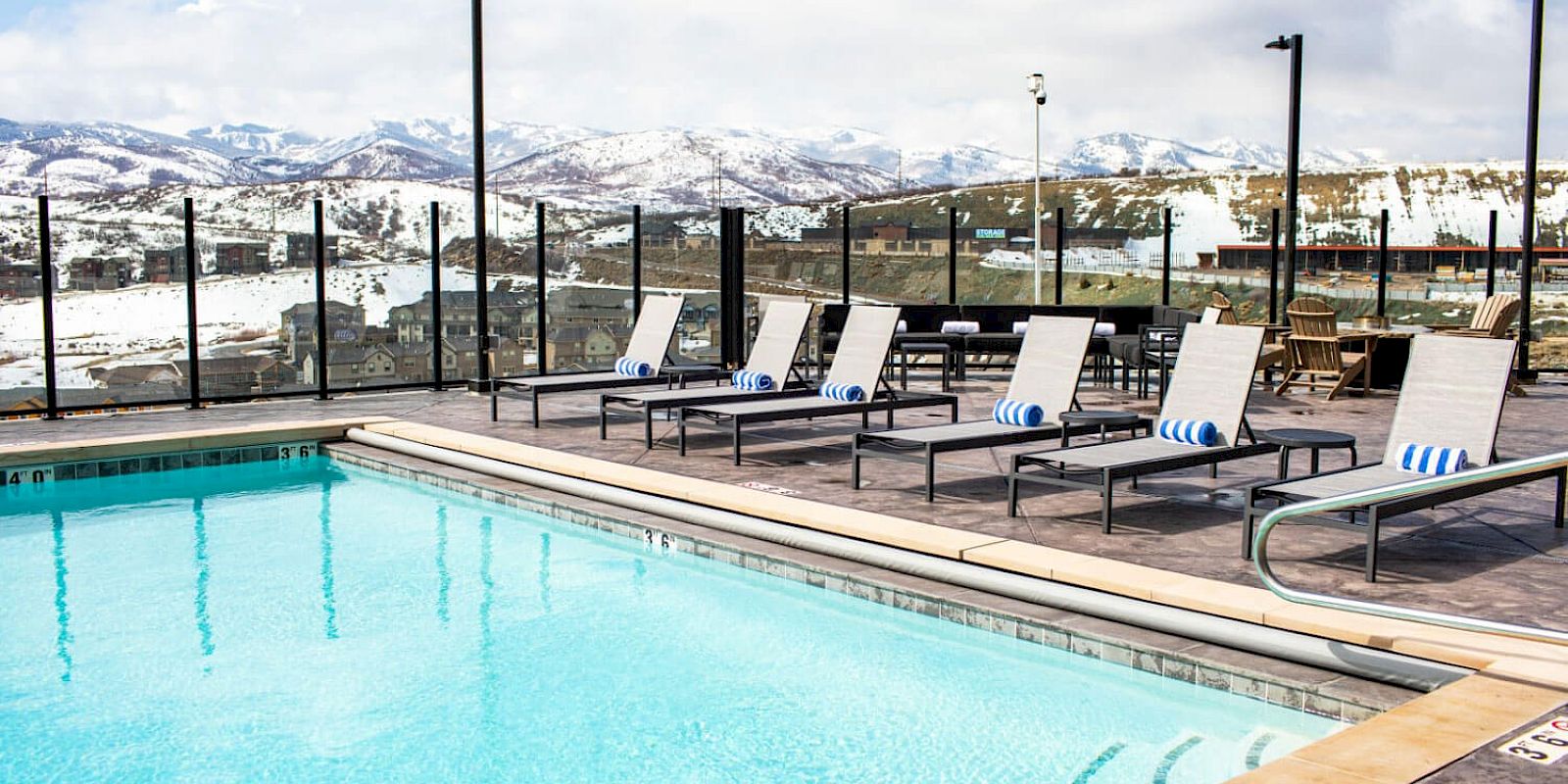 This image shows an outdoor swimming pool with lounge chairs and snowy mountains in the background on a sunny day, surrounded by a fence.