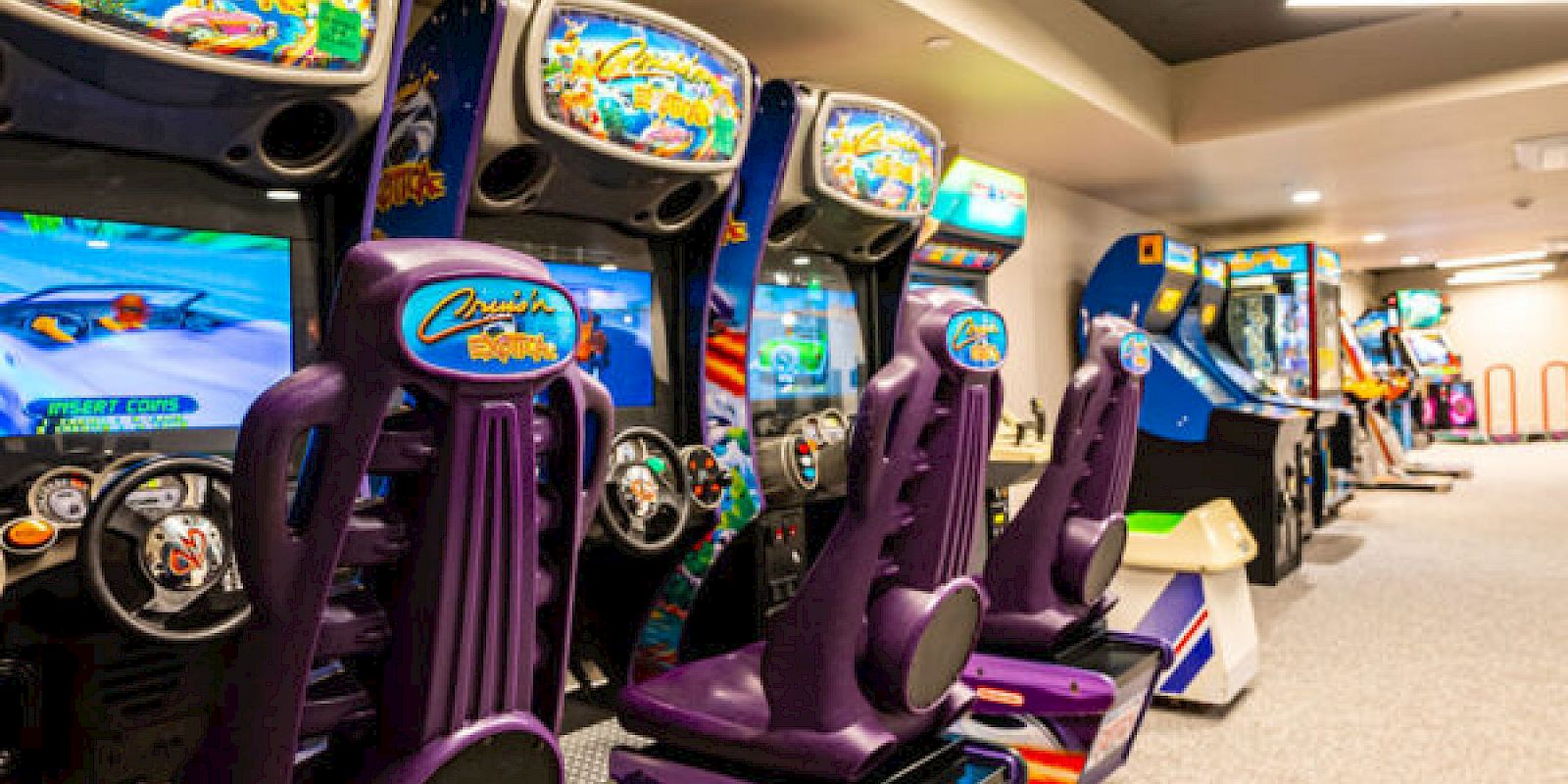 The image shows a row of colorful arcade racing games with purple racing seats and steering wheels in an indoor arcade setting.