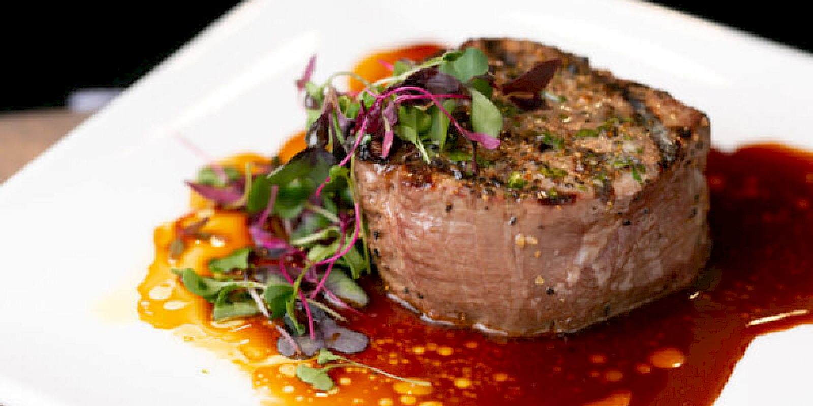 A grilled steak garnished with microgreens, served with sauce on a white plate, ending the sentence.