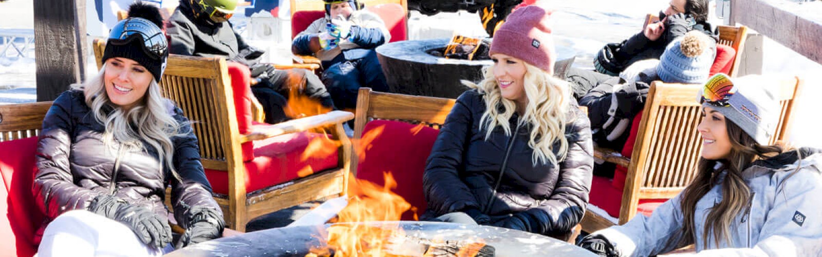 People in winter clothing relax around outdoor fire pits in a snowy setting, with a red truck parked in the background.