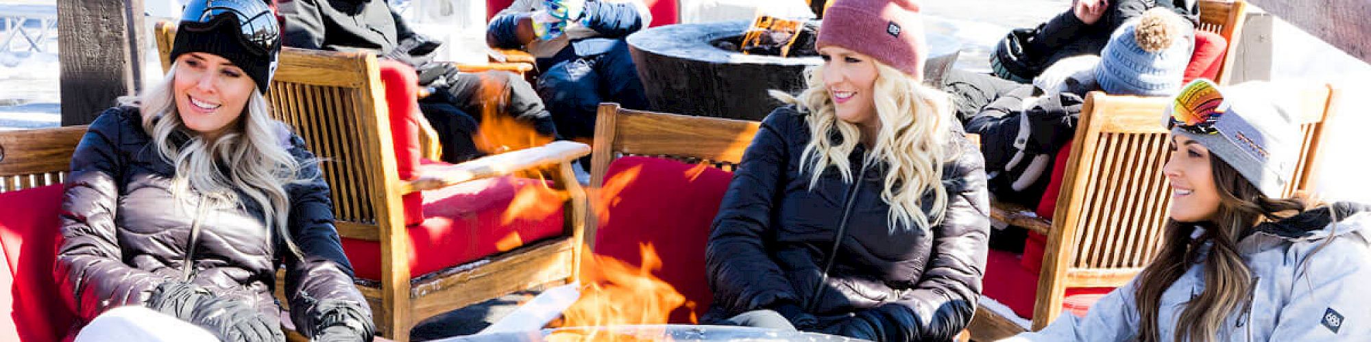 People in winter clothing sit around a fire pit outdoors, with a red truck and buildings in the background.