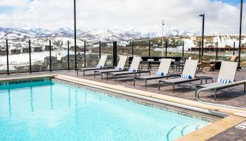 The image shows a swimming pool with lounge chairs and towels, surrounded by a glass fence, overlooking snowy mountains under a partly cloudy sky.