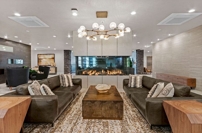 A modern living room with two brown sofas, a central wooden table, a decorative chandelier, and a fireplace under a TV at the far end of the room.