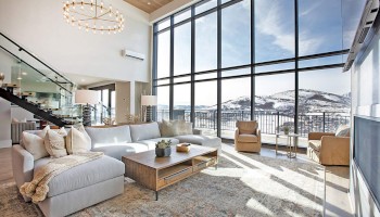 Spacious living room with large windows, a sectional sofa, armchairs, a wooden coffee table, a chandelier, and a scenic mountain view.