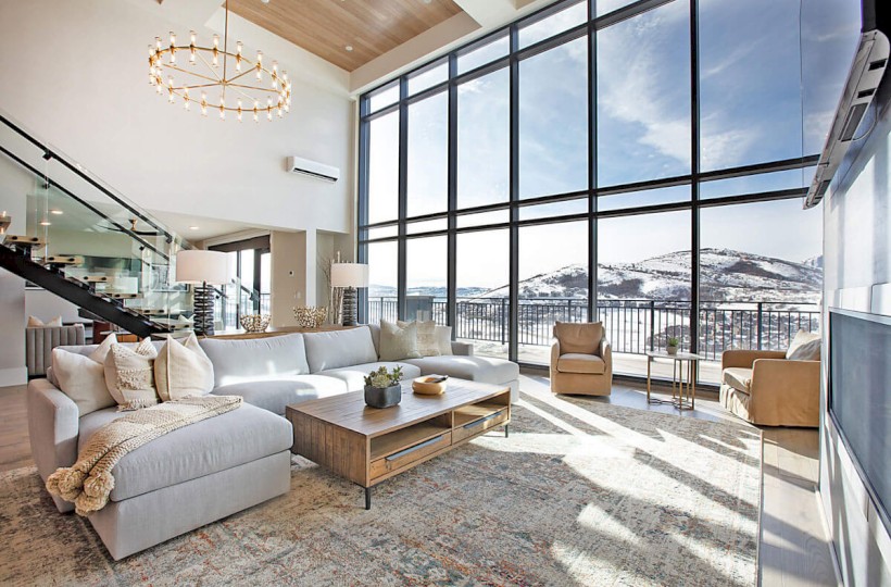 Spacious living room with large windows, a sectional sofa, armchairs, a wooden coffee table, a chandelier, and a scenic mountain view.