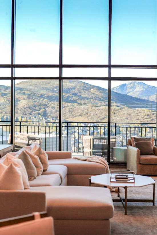 A modern living room with large glass windows overlooking mountains, featuring a beige sofa, armchairs, lamps, and a coffee table.