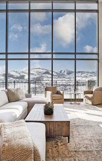 A modern living room with large windows overlooking snowy mountains, a cozy white sofa, a wooden coffee table, and a fireplace on the right.