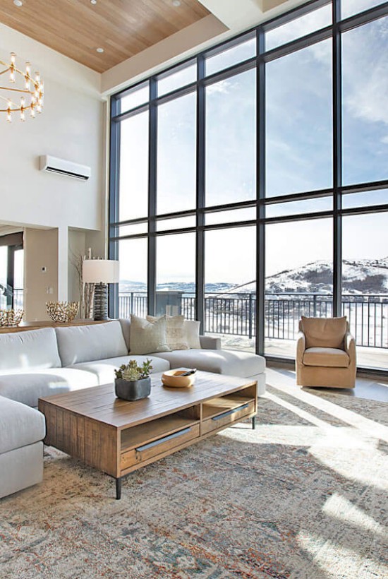 A modern living room with a large sectional sofa, a wooden table, and a stunning mountain view through floor-to-ceiling windows.