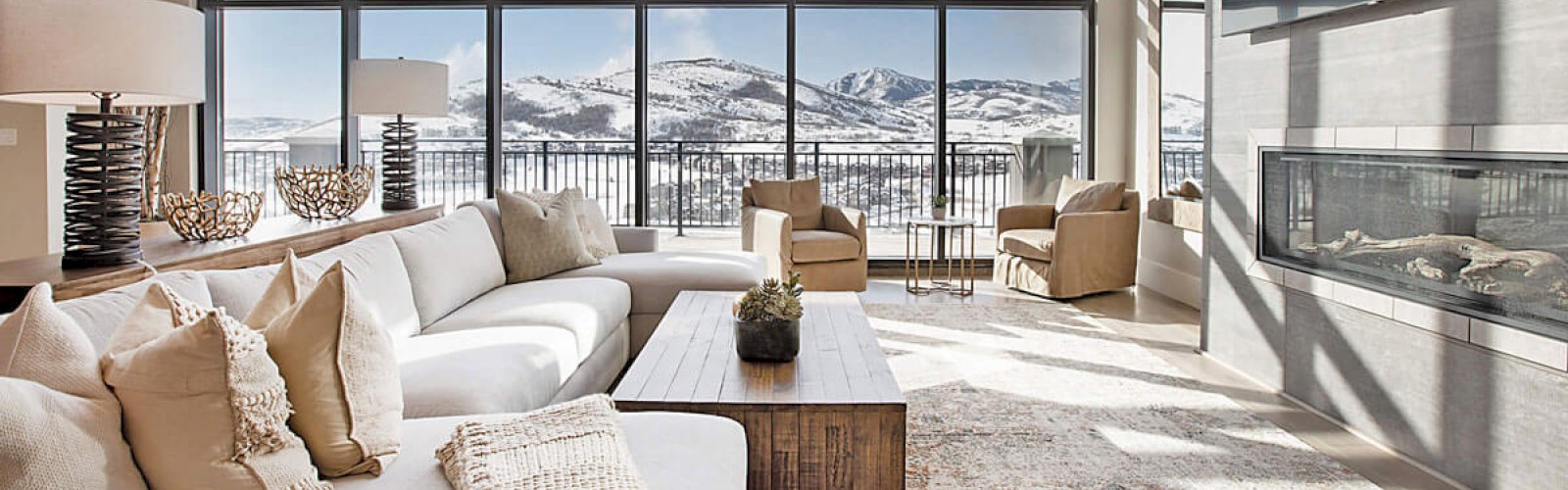 A modern living room with a large sofa, a fireplace, and floor-to-ceiling windows offering a scenic view of snow-covered mountains.