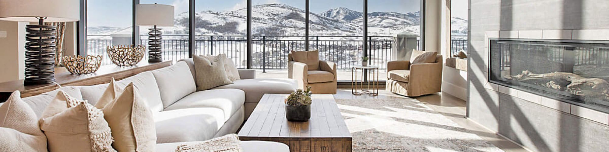 A modern living room with a large sofa, a fireplace, and floor-to-ceiling windows offering a scenic view of snow-covered mountains.