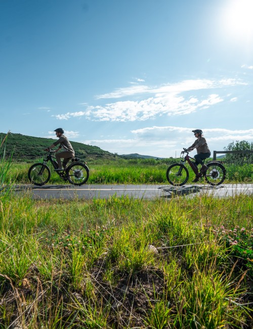 Two people are riding bikes on a sunny, open countryside road surrounded by green fields and hills under a bright blue sky.