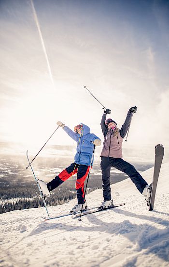 Two people, dressed in winter clothing and ski equipment, joyfully pose with their ski poles raised on a snowy slope under a sunny sky.