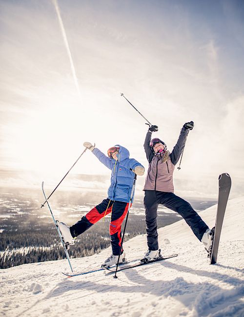 Two people, dressed in winter clothing and ski equipment, joyfully pose with their ski poles raised on a snowy slope under a sunny sky.