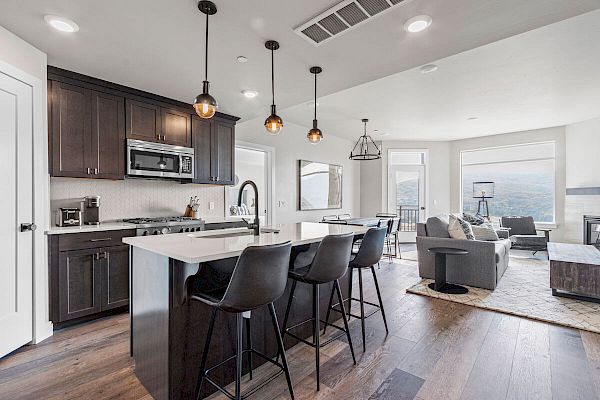 A modern open-concept kitchen and living area with dark cabinets, an island with bar stools, pendant lights, and large windows.